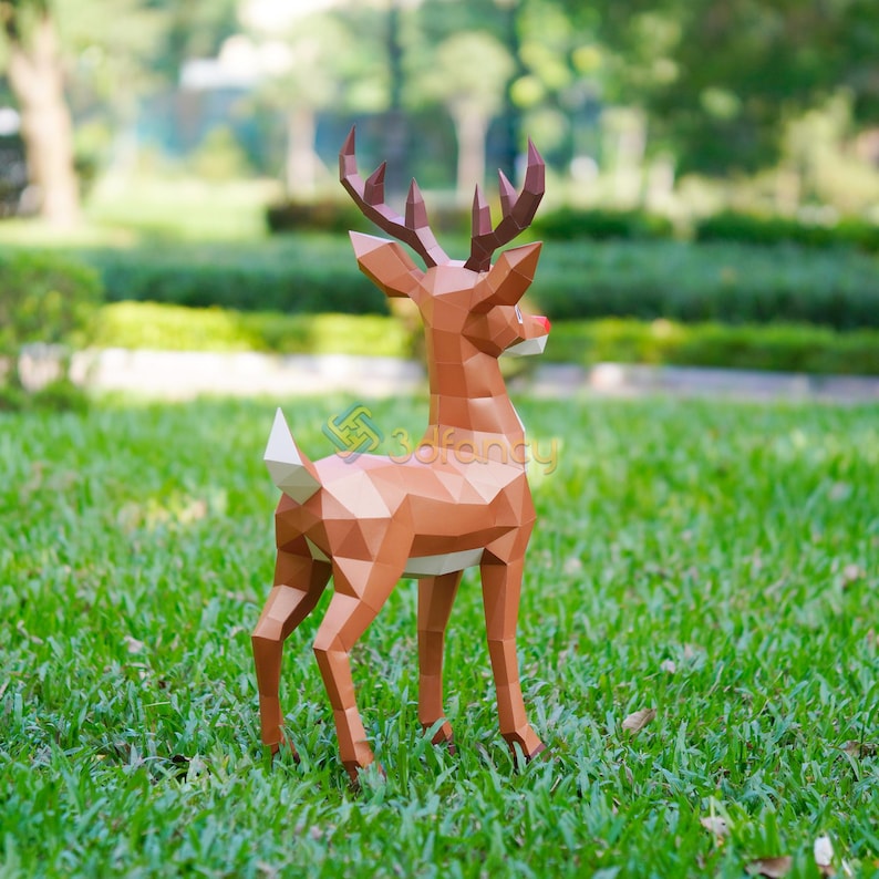 May include: A brown geometric papercraft reindeer with a red nose and large antlers stands on green grass. The reindeer is made of many triangular and polygonal shapes. The background is a blurred green park setting.