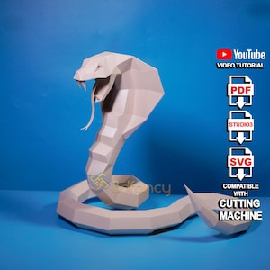 Puede incluir: Un modelo de cobra de papel blanco con su capucha levantada. El modelo está sobre un fondo azul. La imagen también incluye texto que dice "YouTube Video Tutorial", "PDF", "STUDIO3", "SVG", "Compatible con la máquina de corte".