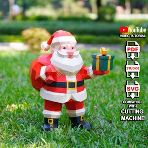 Op de afbeelding: Een papieren Santa Claus-figuur, gekleed in een rood pak met witte randen, een zwarte riem en zwarte laarzen. Hij houdt een groene en gouden geschenkdoos vast. De afbeelding bevat tekst voor een YouTube-videotutorial.