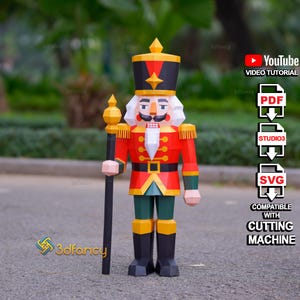 Puede incluir: Una figura de cascanueces de papel geométrico en rojo, verde, negro y dorado, sosteniendo un bastón largo. El diseño presenta un sombrero alto, barba blanca y detalles de uniforme. Texto: YouTube Video Tutorial, PDF, STUDIO3, SVG, Cutting Machine.