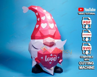 May include: A papercraft gnome with a red hat decorated with white hearts, holding a heart-shaped sign that says "love". The gnome has a white beard and is set against a blue background. The image includes icons for video tutorial, PDF, Studio3, and SVG files.