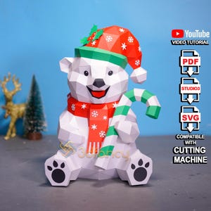 May include: A white papercraft polar bear wearing a red and white Christmas hat and scarf, holding a green and white candy cane. The bear has black paw prints and a smiling face. The image includes text: "YouTube VIDEO TUTORIAL", "PDF", "STUDIO3", "SVG", and "COMPATIBLE WITH CUTTING MACHINE".
