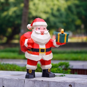 May include: A geometric papercraft Santa Claus figure, wearing a red suit with white trim, a black belt with a gold buckle, and a red hat. He holds a green gift box with a gold ribbon. The figure has a white beard and a red sack.