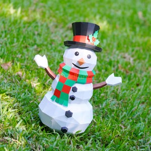 May include: A white paper snowman with a black top hat, a carrot nose, and a red and green scarf. The snowman has black button eyes and a smiling mouth. The arms are brown, and the hands are white. The snowman is standing on green grass.