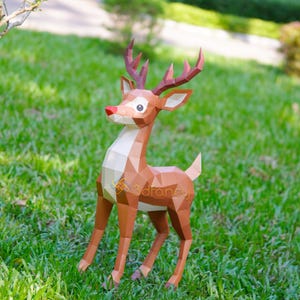 May include: A brown papercraft deer with a red nose and large antlers stands on green grass. The deer has a white chest and geometric design. The logo "3dfancy" is visible on the deer's side. This decorative item is suitable for holiday decor.
