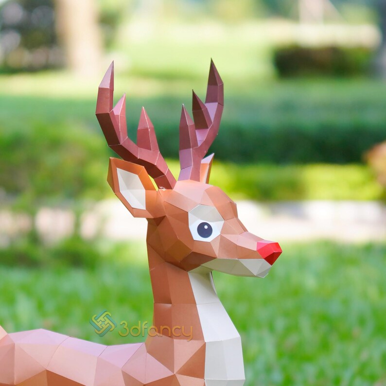 May include: A geometric papercraft reindeer sculpture with brown and tan tones, a red nose, and large, expressive eyes. The antlers are a darker brown. The sculpture is set against a blurred green background, suggesting an outdoor setting.
