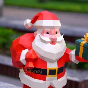 May include: A geometric papercraft Santa Claus figure, featuring a red suit, white beard, and a matching hat. The figure holds a green gift box with a yellow ribbon and a red sack. The logo "3dfancy" is visible on the figure.