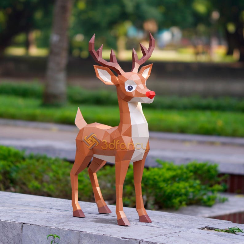 May include: A brown geometric papercraft reindeer with a red nose, white chest, and brown antlers. The reindeer is standing on a concrete surface in front of green foliage. The logo "3dFancy" is visible on the side.