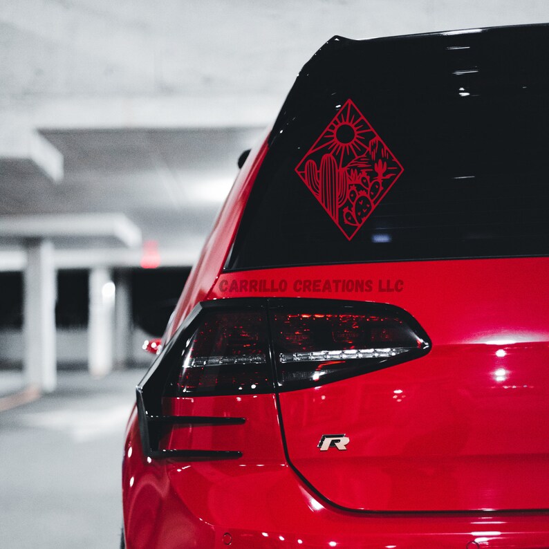 May include: Red car with a black rear window and a red vinyl decal of a desert scene with a cactus, sun, and mountains. The decal is in a diamond shape and says "Carrillo Creations LLC" below it.