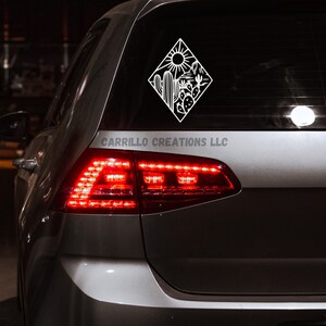 May include: A white vinyl decal of a desert scene with a sun, cacti, and mountains. The decal is on the back window of a silver car. The text "CARRILLO CREATIONS LLC" is visible below the decal.
