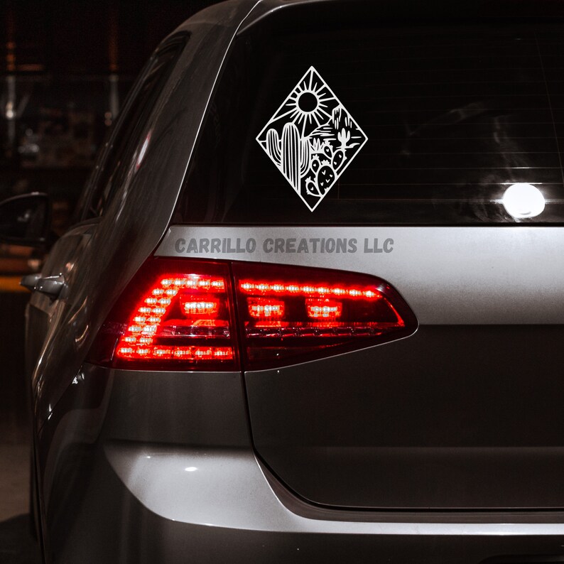 May include: A white vinyl decal of a desert scene with a sun, cacti, and mountains. The decal is on the back window of a silver car. The text "CARRILLO CREATIONS LLC" is visible below the decal.