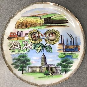 May include: A decorative plate with a gold rim featuring colorful illustrations of Kansas landmarks. The plate includes images of a wheat field, sunflowers, cows, an industrial scene, and the Kansas State Capitol building. Text on the plate reads "Kansas" and other descriptions.