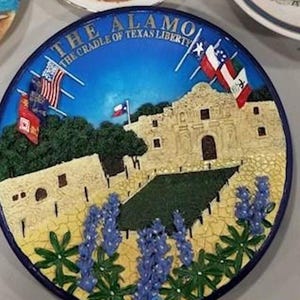 May include: A decorative plate featuring a depiction of The Alamo, with flags and the text "THE ALAMO THE CRADLE OF TEXAS LIBERTY." The plate has a blue border, a blue sky background, and includes images of bluebonnets.