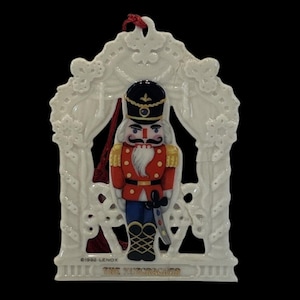 May include: A white porcelain Christmas ornament featuring a nutcracker soldier in a red coat, black hat, and blue pants. The ornament is framed by a white archway with the text "THE NUTCRACKER" at the bottom. A red ribbon is attached for hanging.