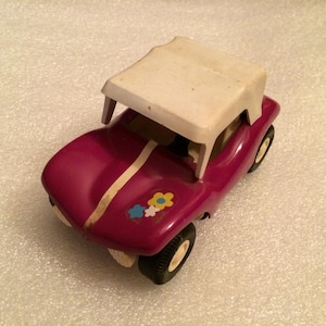 May include: A vintage toy car in a deep magenta hue, featuring a white roof and a white stripe down the center. The car has black tires with cream-colored rims and a floral decal. This classic toy car is a nostalgic collectible.