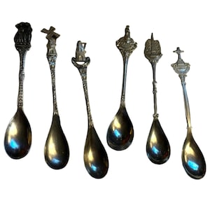 May include: Six vintage silver spoons with ornate handles. Each spoon features a unique design, including a lion, windmill, building, and cross. The spoons have a shiny, reflective surface and are arranged in a row.