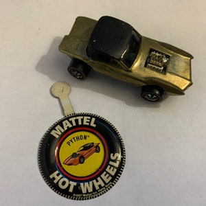 May include: A gold Hot Wheels toy car with a black roof and a visible engine. The car is next to a round, black and yellow button with the words "Mattel", "Python", and "Hot Wheels" on it.