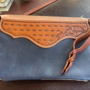 May include: A dark blue leather clutch with a brown leather flap featuring a floral design and colorful accents. The clutch has a long brown leather strap.