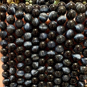 Può includere: Una collana di perle di pietre preziose blu scuro e nere. Le perle sono rotonde e hanno una finitura liscia e lucida. Le perle sono infilate su un filo elastico trasparente.