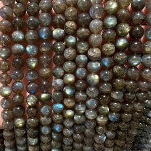 Op de afbeelding: Een streng donkergrijze labradorit edelsteen kralen met iriserende blauwe en groene flitsen. De kralen zijn rond en glad en zijn geregen op een doorzichtige draad.