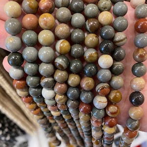 May include: A strand of multi-coloured gemstone beads. The beads are round and have a variety of brown, grey, and red tones. The beads are strung on a wire and are ready to be used for jewellery making.