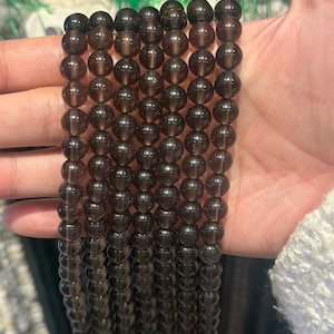 Könnte beinhalten: Eine Strähne aus dunkelbraunen Edelsteinperlen. Die Perlen sind rund und glatt und auf einem durchsichtigen Faden aufgefädelt. Die Perlen haben einen Durchmesser von etwa 8 mm.