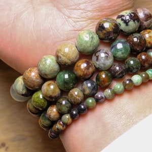 May include: A set of five bracelets made with green, brown, and black gemstone beads. The bracelets are stacked on a person's wrist.