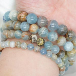 Peut inclure: Plusieurs bracelets de perles bleues, marron et blanches sont empilés sur un poignet. Les bracelets sont composés de perles rondes et polies de différentes tailles. Le fond est un tissu blanc et soyeux.