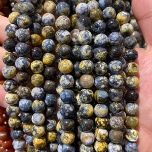 Könnte beinhalten: Stränge aus runden Edelsteinperlen in Schwarz-, Gelb- und Blautönen. Die Perlen haben ein gesprenkeltes Muster und sind aneinandergereiht, wahrscheinlich für die Schmuckherstellung. Die Perlen haben einen Durchmesser von etwa 8 mm.