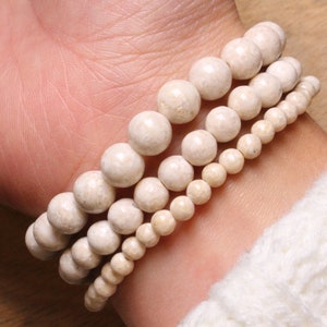 Puede incluir: Tres pulseras de cuentas blancas apiladas en una muñeca. Las pulseras están hechas de cuentas redondas y lisas de diferentes tamaños.
