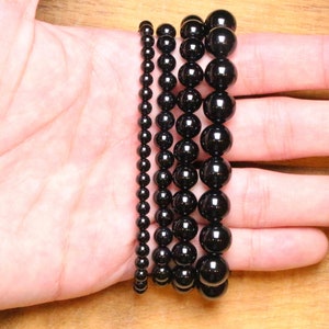 May include: Five black beaded bracelets of varying sizes, stacked on a person's hand.