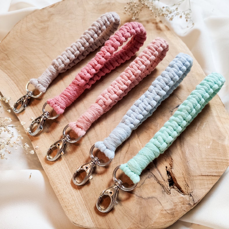 May include: Five colourful macrame keychains with silver key rings. The keychains are made with a variety of colours, including pink, blue, and green.