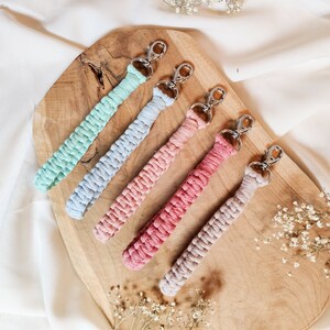 May include: Five colourful braided keychains with silver key rings. The keychains are in shades of blue, pink, and beige. The keychains are displayed on a wooden surface.
