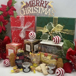 May include: A Christmas gift hamper containing two wrapped boxes, a box of breakfast tea, a fruit cake, shortbread fingers, two jars of preserves, two gingerbread cookies, and several wrapped candies, decorated with red poinsettias and a Merry Christmas sign.