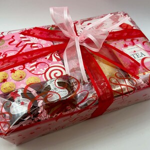 May include: A rectangular gift basket wrapped in clear cellophane with a red and pink patterned design. The basket contains Reids Candy Cane shortbread, a box of Afternoon Tea, and two small jars of preserves, all tied with a pink ribbon.