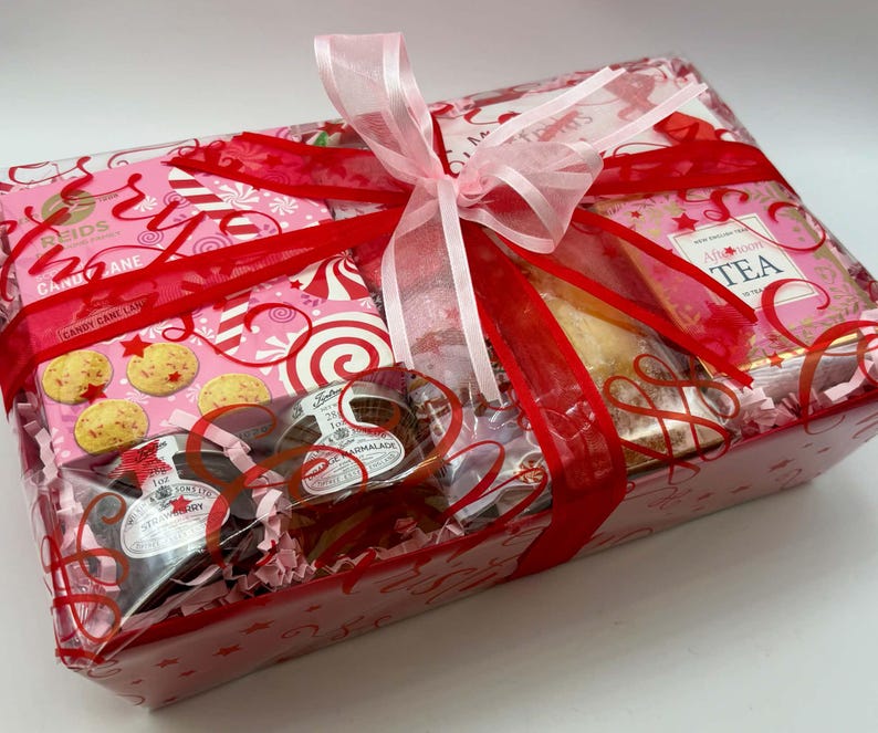 May include: A rectangular gift basket wrapped in clear cellophane with a red and pink patterned design. The basket contains Reids Candy Cane shortbread, a box of Afternoon Tea, and two small jars of preserves, all tied with a pink ribbon.