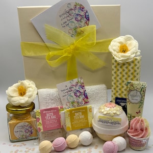 May include: A cream-coloured gift set tied with a yellow ribbon, containing bath products. Includes bath bombs, bath tea bags, a jar of bath fizz, a hand cream tube, and decorative flowers. The set is decorated with confetti.