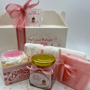 Puede incluir: Una caja de regalo blanca con una cinta rosa y una etiqueta rosa que dice "Have A Happy Bath Day". La caja contiene una bomba de ba&ntilde;o rosa con forma de cupcake, un frasco de sales de ba&ntilde;o, una pastilla de jab&oacute;n rosa y una toalla blanca. La caja est&aacute; rodeada de confeti rosa y blanco.
