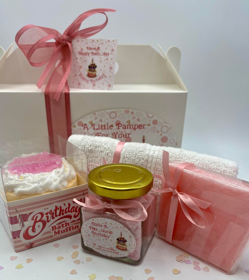 Puede incluir: Una caja de regalo blanca con una cinta rosa y una etiqueta rosa que dice "Have A Happy Bath Day". La caja contiene una bomba de ba&ntilde;o rosa con forma de cupcake, un frasco de sales de ba&ntilde;o, una pastilla de jab&oacute;n rosa y una toalla blanca. La caja est&aacute; rodeada de confeti rosa y blanco.