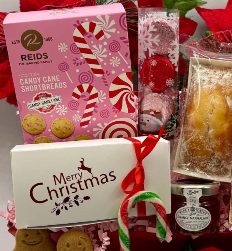May include: A holiday gift set featuring a pink box of Reids Scottish Candy Cane Shortbreads, a clear tube of three foil-wrapped chocolates, a white box labeled Merry Christmas with a red ribbon and candy cane, and a jar of orange marmalade.