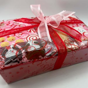 May include: A rectangular gift basket wrapped in clear cellophane with a large pink and red ribbon. Inside are boxes of Reids cookies, jars of strawberry jam and marmalade, and a box of tea, all nestled on pink shredded paper filler.