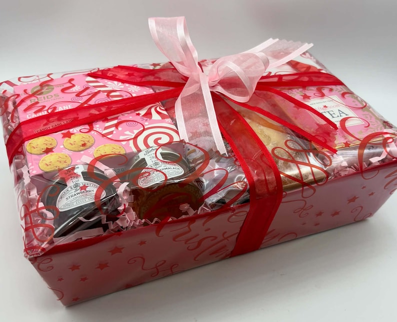 May include: A rectangular gift basket wrapped in clear cellophane with a large pink and red ribbon. Inside are boxes of Reids cookies, jars of strawberry jam and marmalade, and a box of tea, all nestled on pink shredded paper filler.