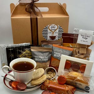 May include: A gift box labeled Your Little Coffee Shop with a brown ribbon, containing various snacks including Grandma Wild's biscuits, Rosie & Roni's fudge, a Madeira cake, and a teacup filled with dark tea on a saucer with small chocolates.