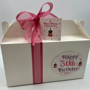 Puede incluir: Caja de regalo blanca con una cinta rosa y una etiqueta que dice "A Happy Birthday Treat For You". La caja tiene una pegatina en la parte delantera que dice "Happy 50th Birthday" con un coraz&oacute;n rosa y dos ilustraciones de pasteles.
