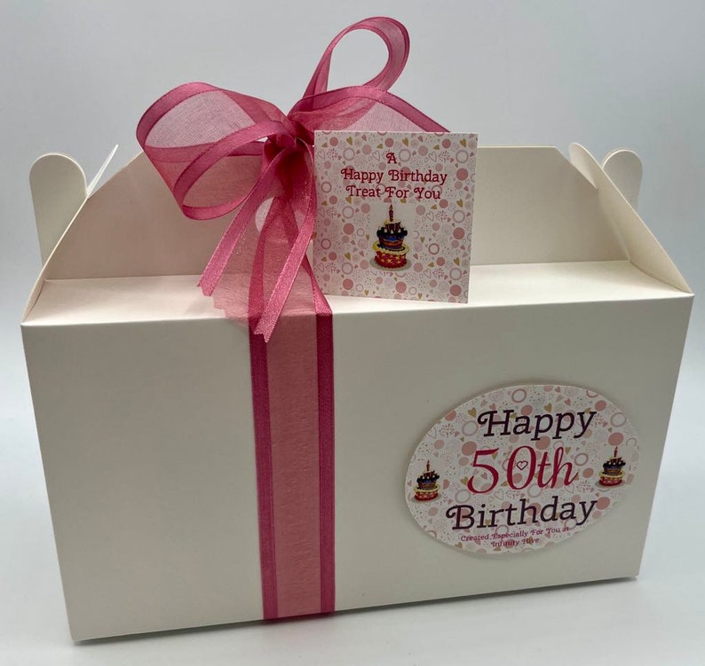 Puede incluir: Caja de regalo blanca con una cinta rosa y una etiqueta que dice "A Happy Birthday Treat For You". La caja tiene una pegatina en la parte delantera que dice "Happy 50th Birthday" con un coraz&oacute;n rosa y dos ilustraciones de pasteles.