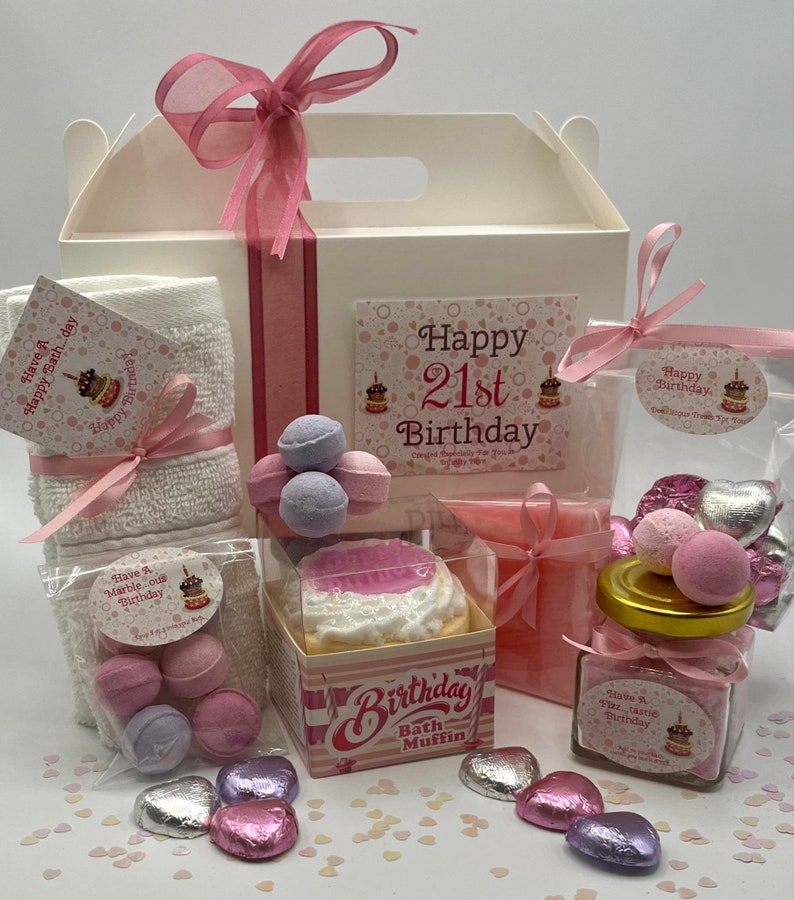 Puede incluir: Una caja de regalo blanca llena de productos de ba&ntilde;o rosas y blancos para un 21 cumplea&ntilde;os. La caja incluye una toalla de ba&ntilde;o, bombas de ba&ntilde;o, sales de ba&ntilde;o, un muffin de ba&ntilde;o y una pastilla de jab&oacute;n. La caja est&aacute; decorada con una cinta rosa y una tarjeta que dice "Feliz 21 cumplea&ntilde;os".