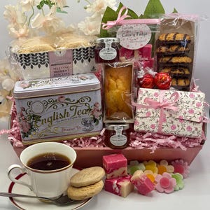 May include: A gift basket containing a tin of English Tea, a box of Melting Moment biscuits, a loaf cake, jars of strawberry jam and orange marmalade, chocolate-covered cookies, Lindor truffles, and fudge, with a teacup and saucer in front.