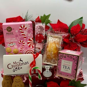 May include: A Christmas gift hamper containing Reids candy cane shortbread, a loaf cake, New English Tea, two jars of jam, gingerbread cookies, foil-wrapped chocolates, and a candy cane, accented with red poinsettia flowers and a Merry Christmas sign.