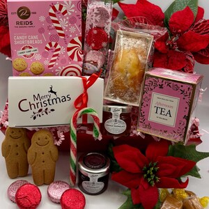 May include: A holiday gift hamper containing Reids Candy Cane Shortbreads, a Merry Christmas box, New English Tea, two gingerbread cookies, two jars of preserves, a candy cane, wrapped chocolates, and red poinsettia floral decorations.
