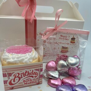 Puede incluir: Una caja de regalo blanca con una cinta rosa y una bolsa de pl&aacute;stico transparente llena de chocolates en forma de coraz&oacute;n rosa, plateado y morado. La caja tambi&eacute;n contiene una bomba de ba&ntilde;o rosa y blanca con forma de cupcake con las palabras "Birthday Bath Muffin" en la parte delantera.